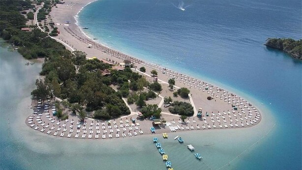 An aerial view of a coastal resort with a circular beach area, multiple rows of beach umbrellas, and a marina with boats. - Karbel Beach Hotel (Fethiye)