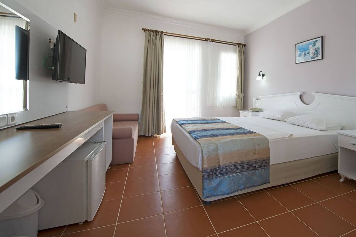 A hotel room with a bed, bedside table, a desk with a chair, a television mounted on the wall, and a window with curtains.