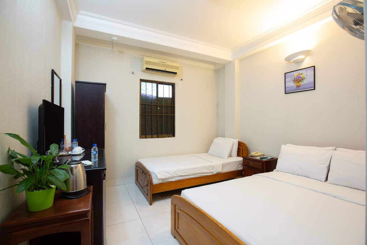 A hotel room with two beds, a wooden dresser, a potted plant, a window with bars, an air conditioning unit, and a bedside table with a lamp.