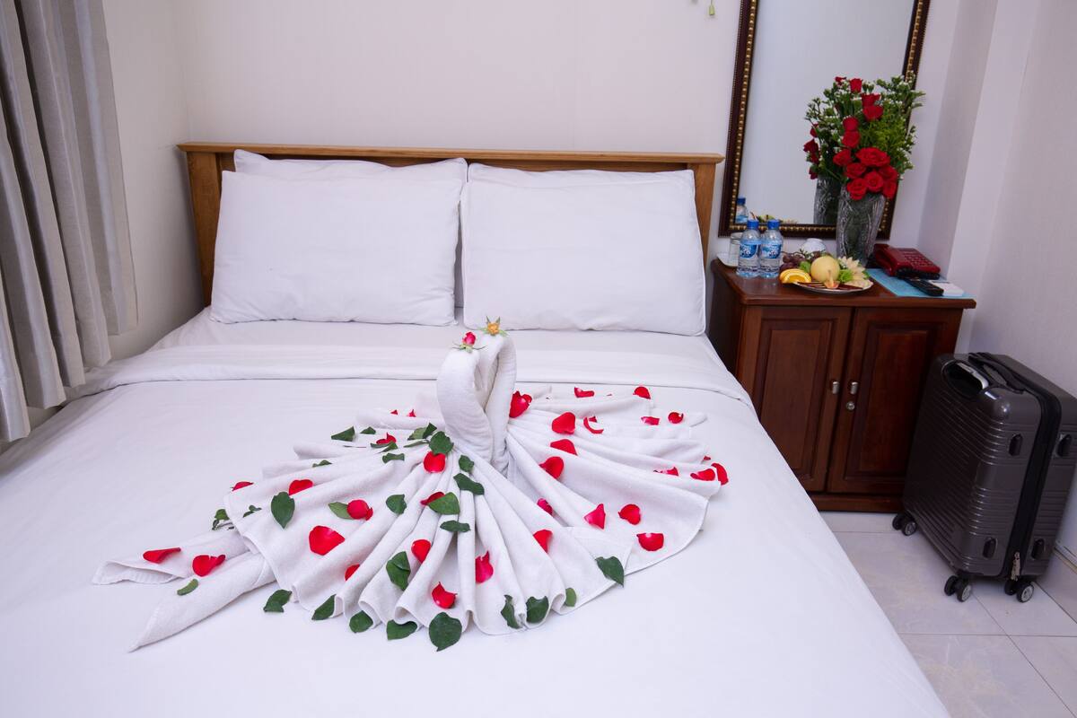 A neatly made bed with a white bedspread and pillows, a wooden headboard, a wooden nightstand with a vase of red roses, a suitcase, and a mirror on the wall.