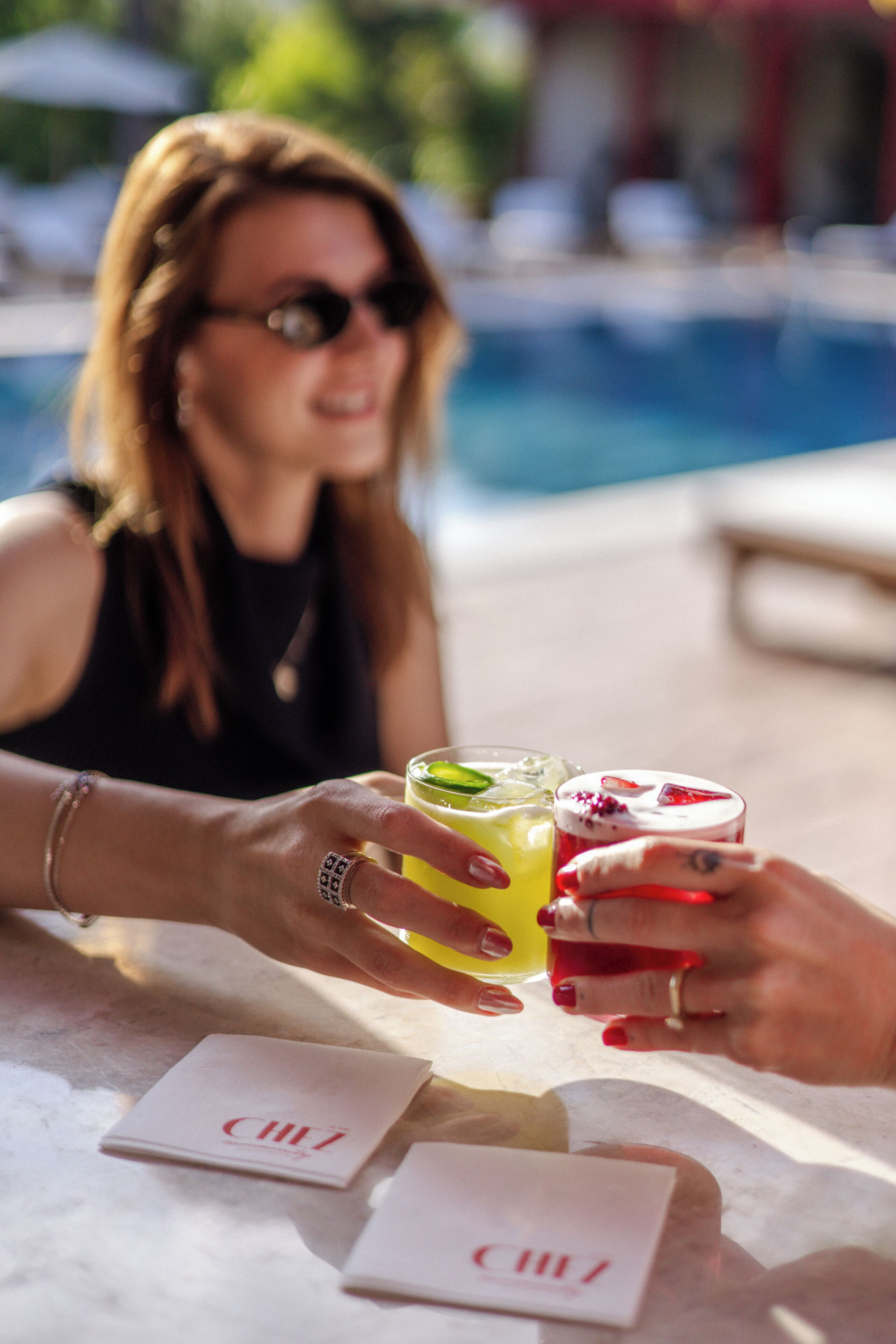 En person som håller i två drinkar vid ett bord vid poolkanten, med en pool i bakgrunden.