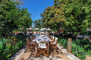A covered outdoor dining area with a long table and chairs, surrounded by lush greenery and a clear blue sky.