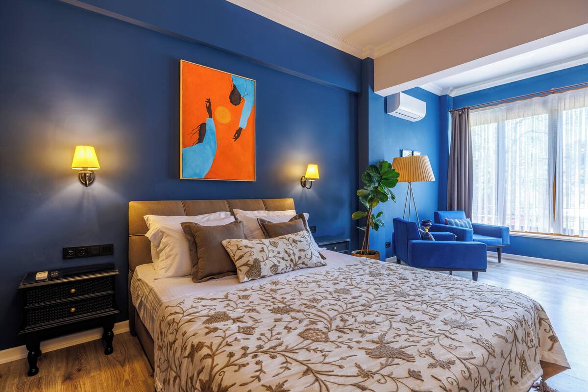 A bedroom with a large bed, two bedside lamps, a nightstand, a potted plant, a blue armchair, and a window with curtains.