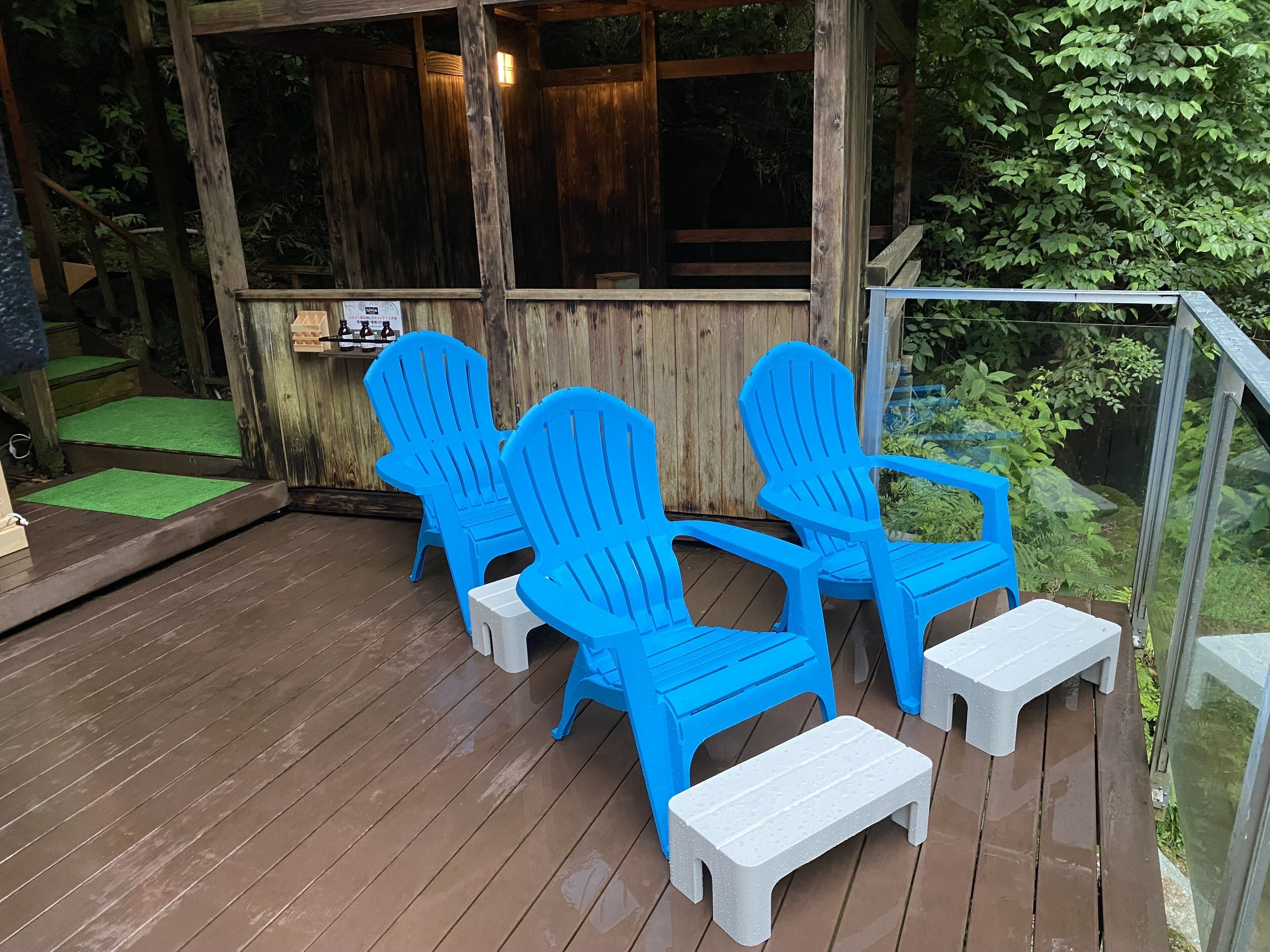 Een houten terras met blauwe Adirondack-stoelen en witte krukken, een glazen omheining en een houten schutting.