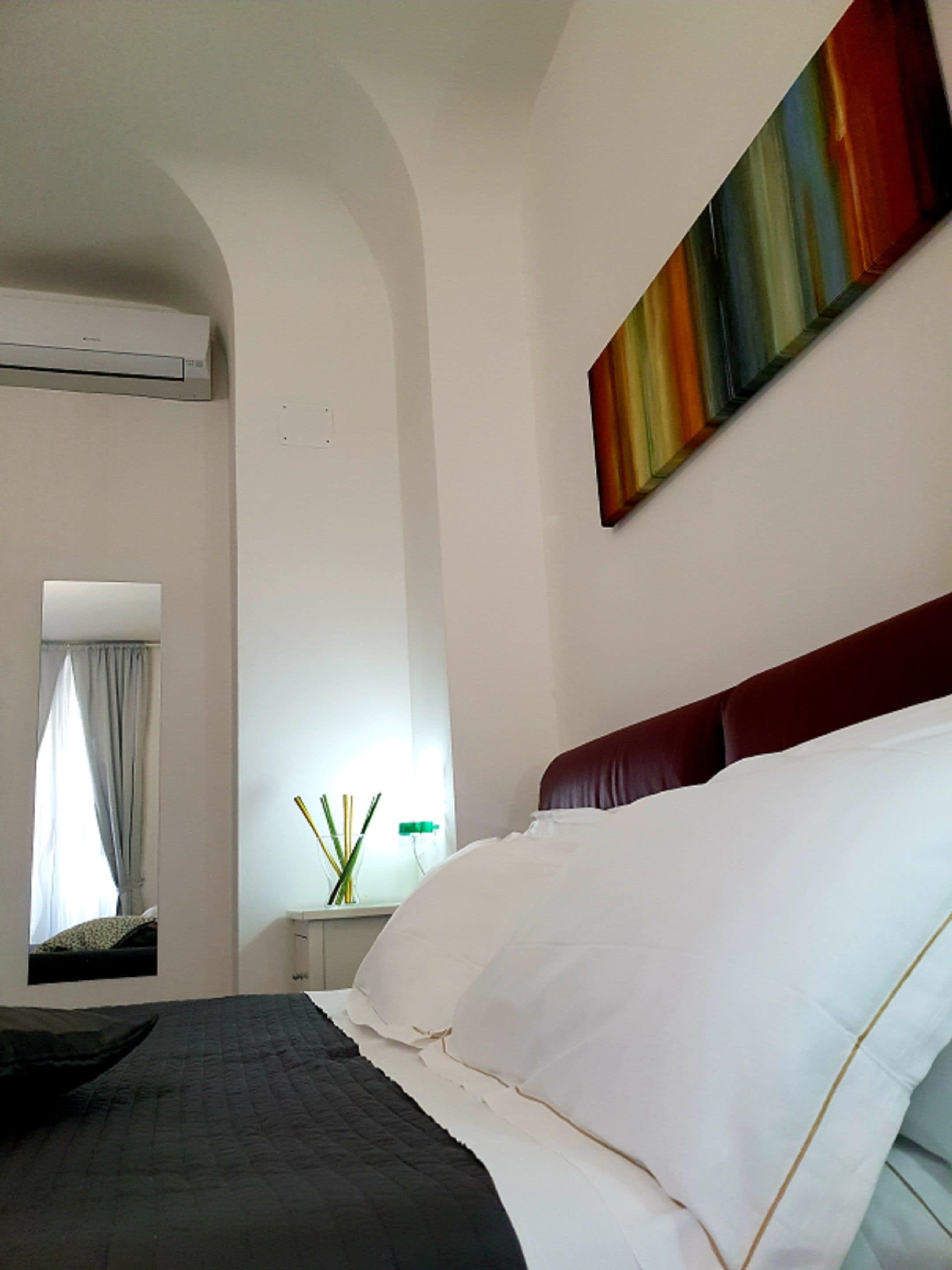 A hotel room with a bed, a window with curtains, an air conditioning unit, a nightstand with a lamp, and a colorful painting on the wall.