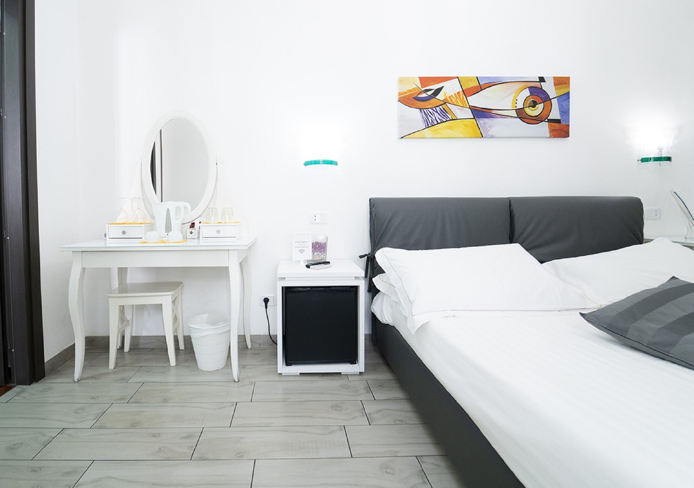 A bedroom with a bed, a desk with a mirror, a small bedside table, and a wall-mounted abstract painting.