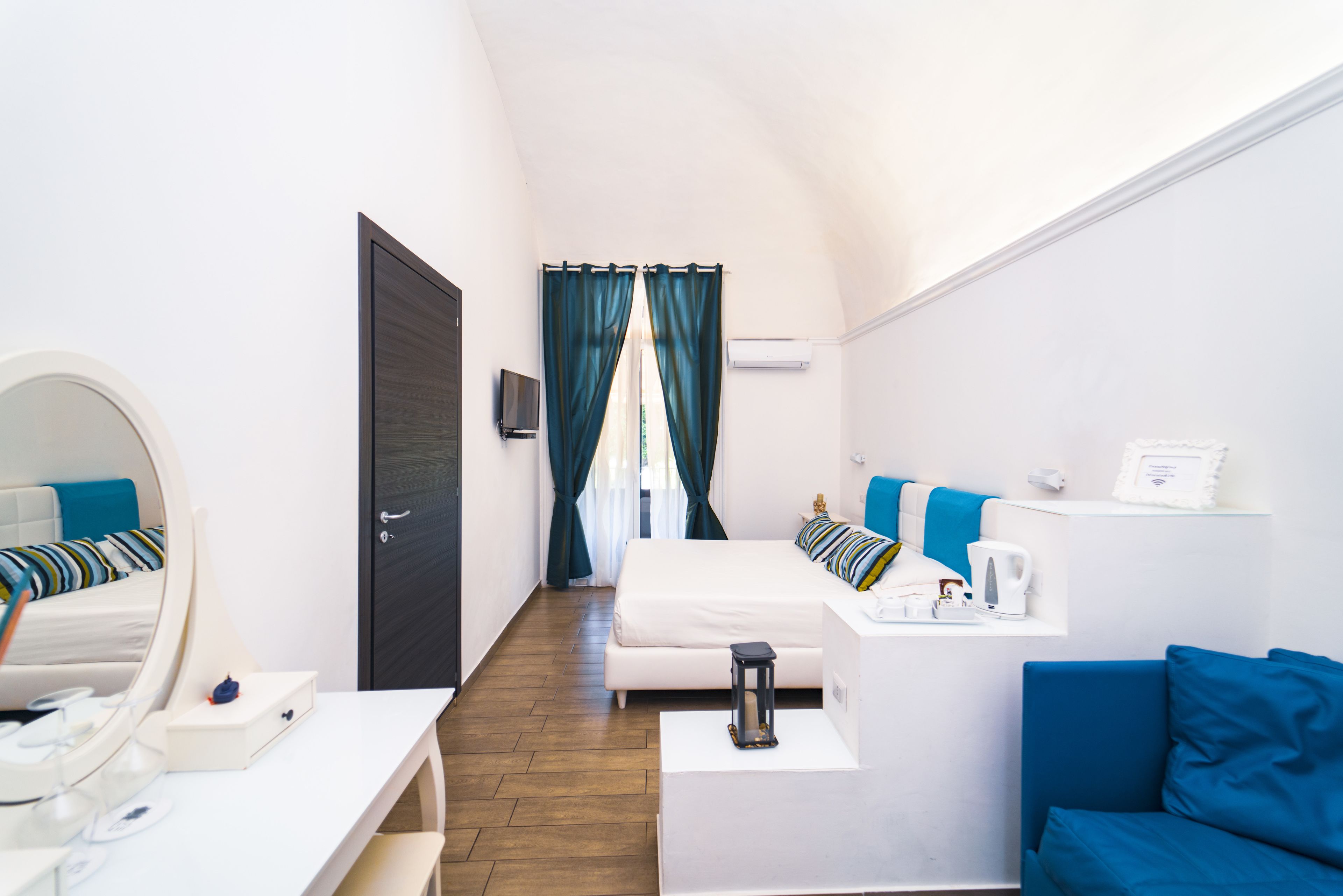 A hotel room with a bed, a desk, a chair, a small table, a mirror, a TV, a window with curtains, and a blue sofa.