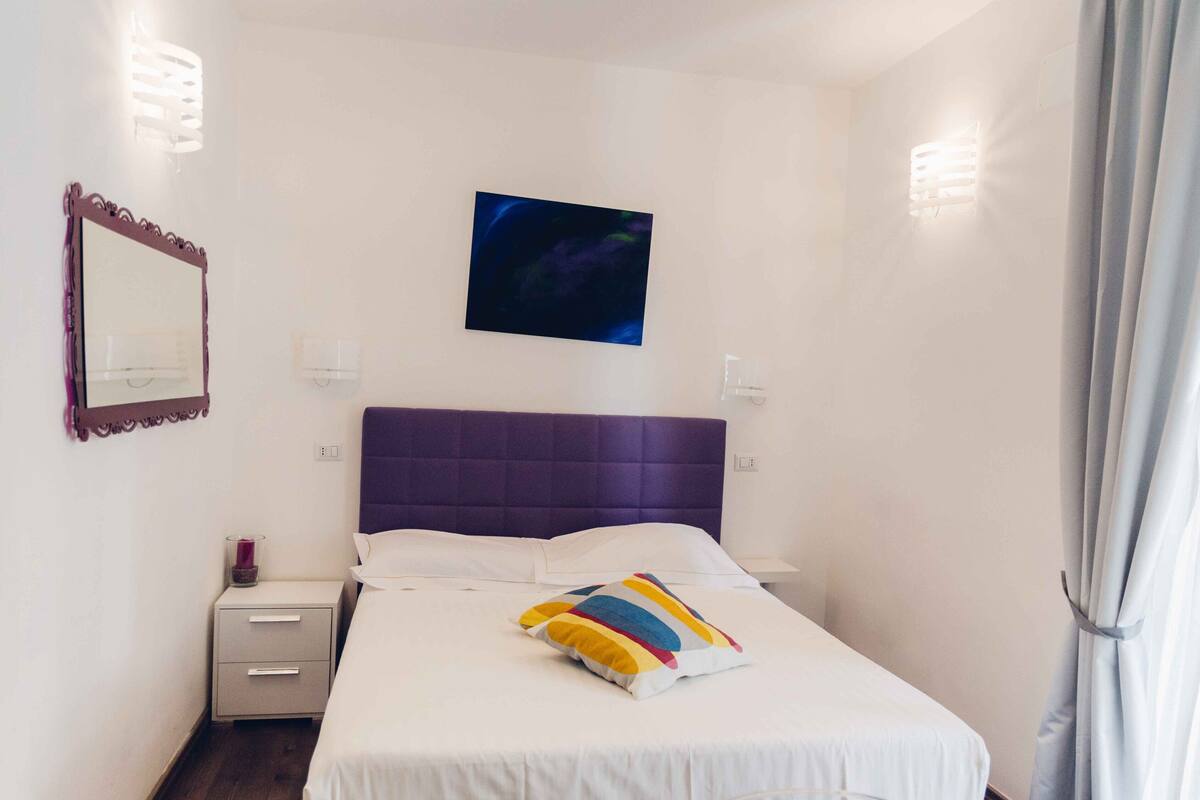 Una camera da letto con una testiera viola, un letto con lenzuola bianche, un cuscino colorato, un comodino con una lampada e un televisore montato a parete.