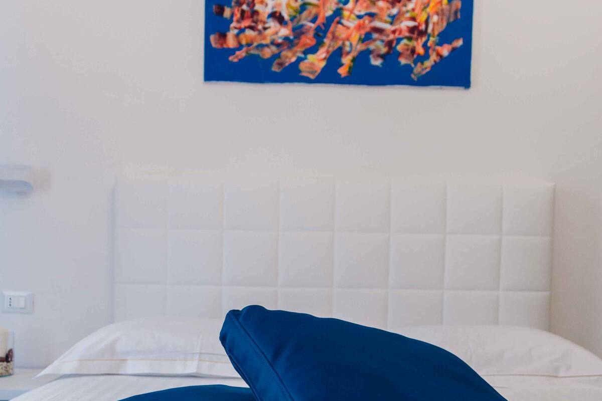 Un letto con biancheria bianca e due cuscini blu, una testiera bianca e un quadro astratto dai colori vivaci appeso alla parete.