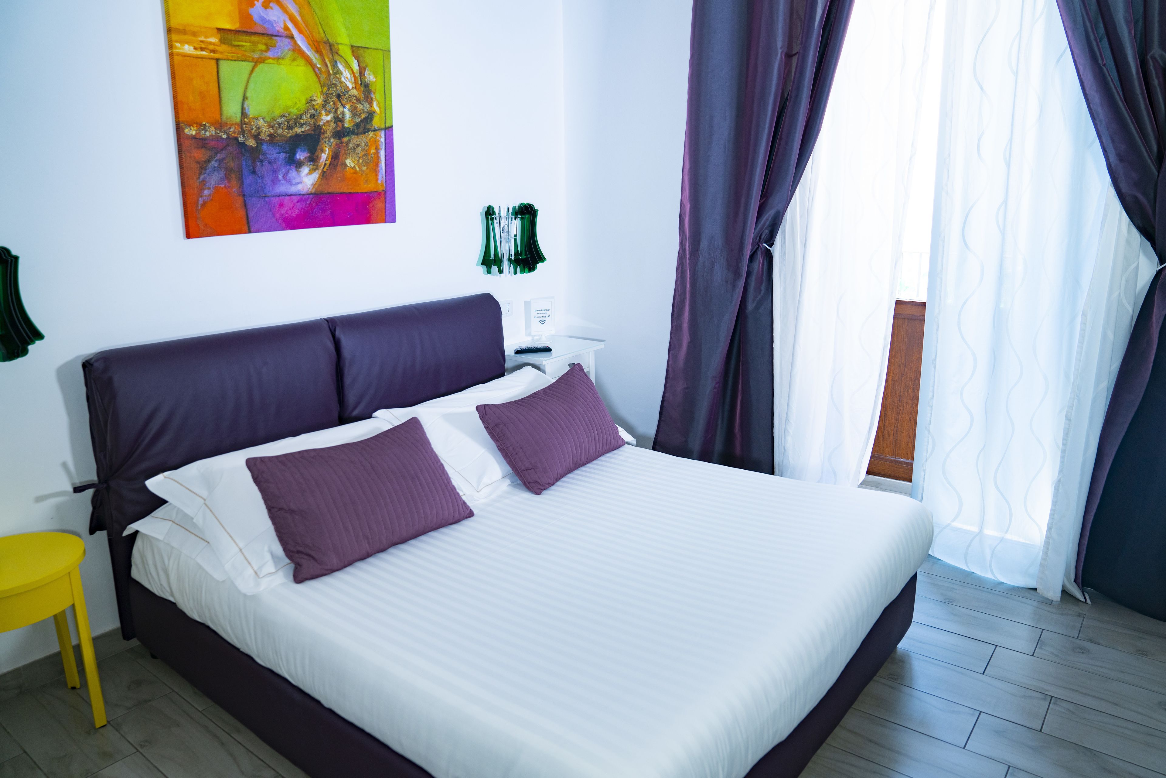 Una cama bien tendida con ropa de cama blanca y morada, un cabecero morado y un cuadro abstracto colorido en la pared.
