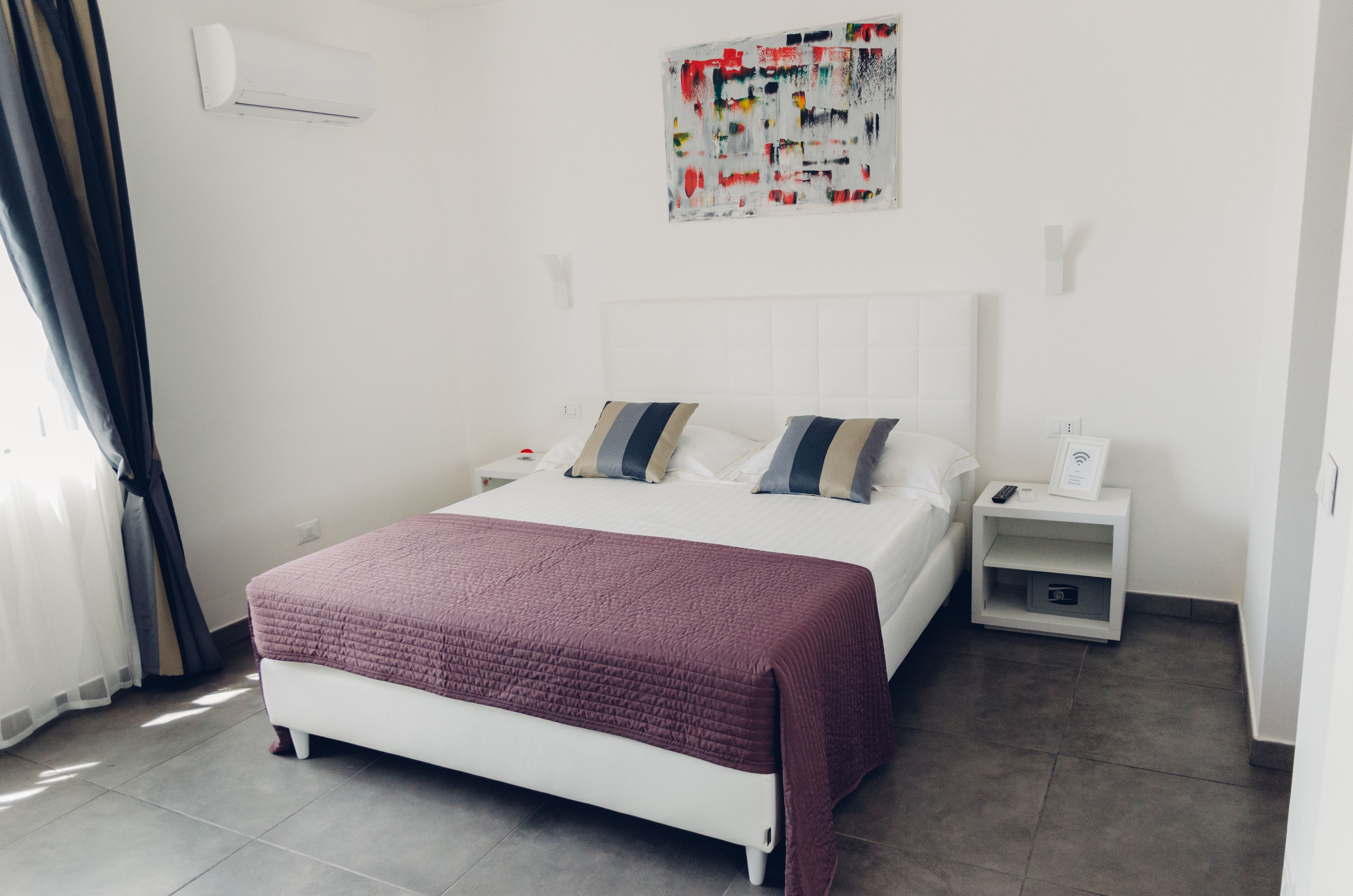 A modern bedroom with a white bed, two pillows, a red blanket, a nightstand with a remote, and a wall art piece.