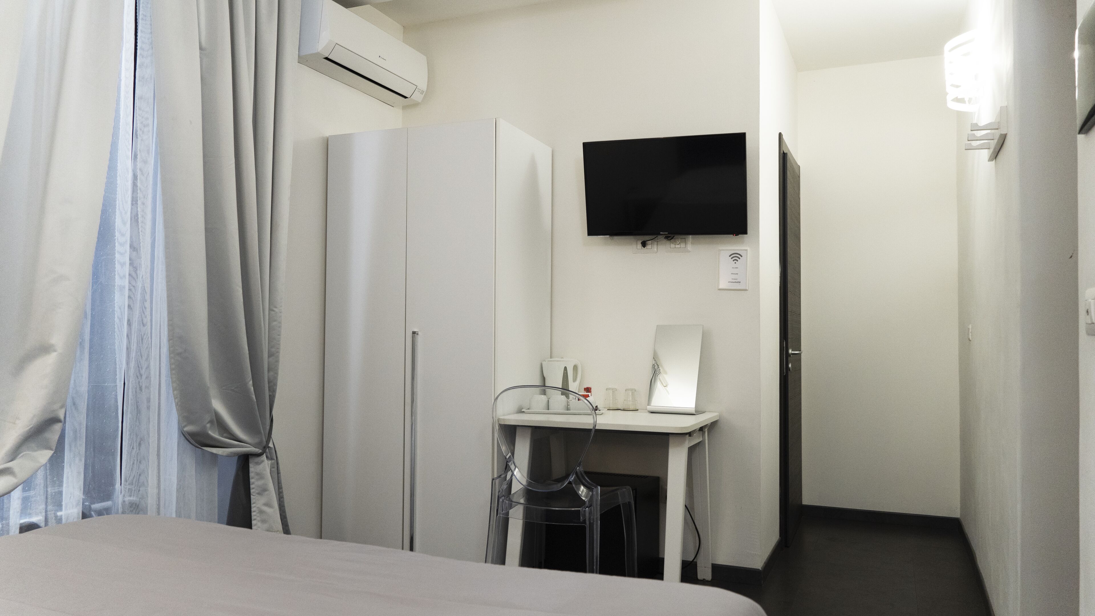 A hotel room with a bed, a desk with a computer, a flat-screen TV, an air conditioning unit, and a door leading to another room.