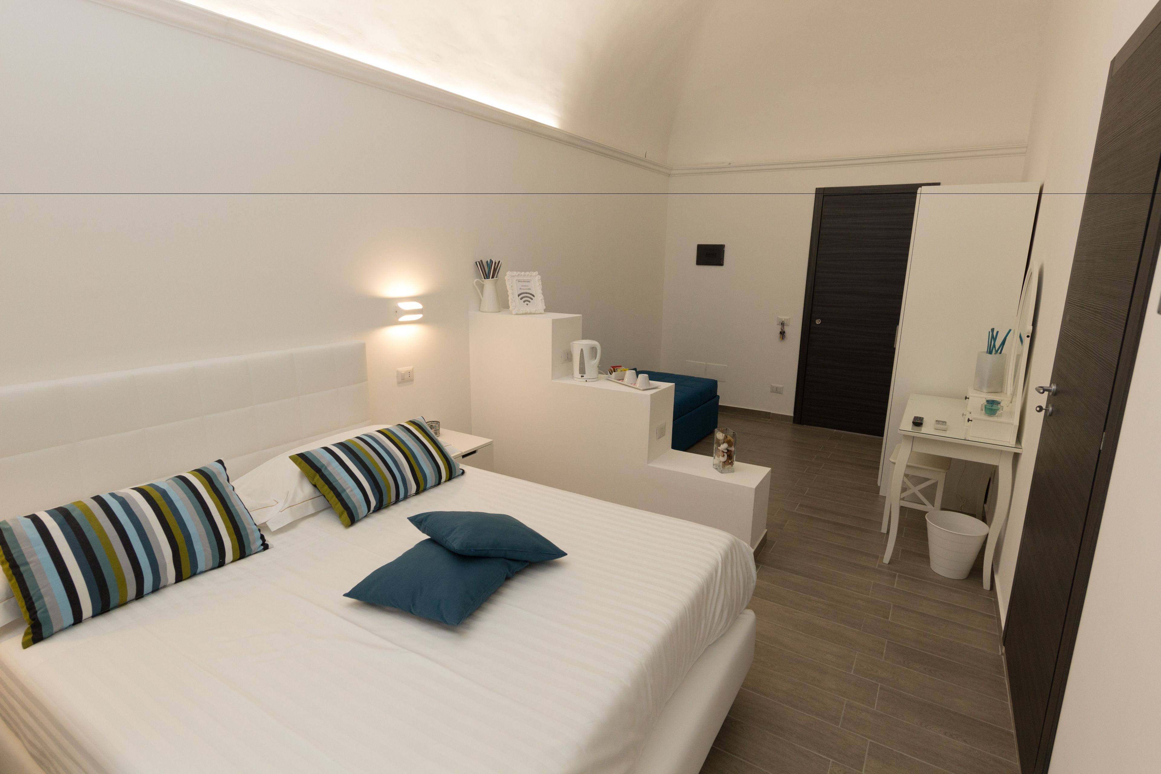 A modern hotel room with a large bed, a bedside table, a desk with a chair, a mirror, and a small trash bin.