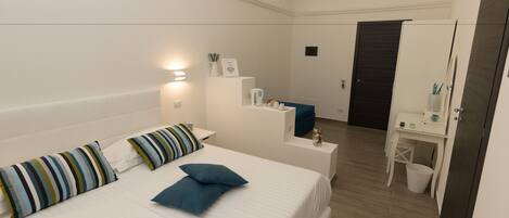 A modern hotel room with a large bed, a bedside table, a desk with a chair, a mirror, and a small trash bin.