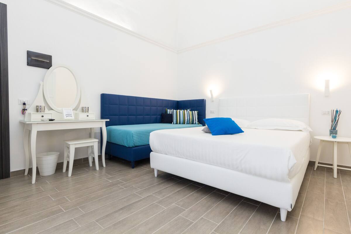 Una camera da letto con un letto bianco, una testiera blu, una scrivania bianca con uno specchio e un piccolo tavolino con una lampada.