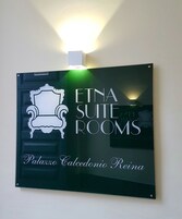 Señalización para 'ETNA SUITE ROOMS' con silueta de una silla y el texto 'Palazzo Calcedonio Regina'.