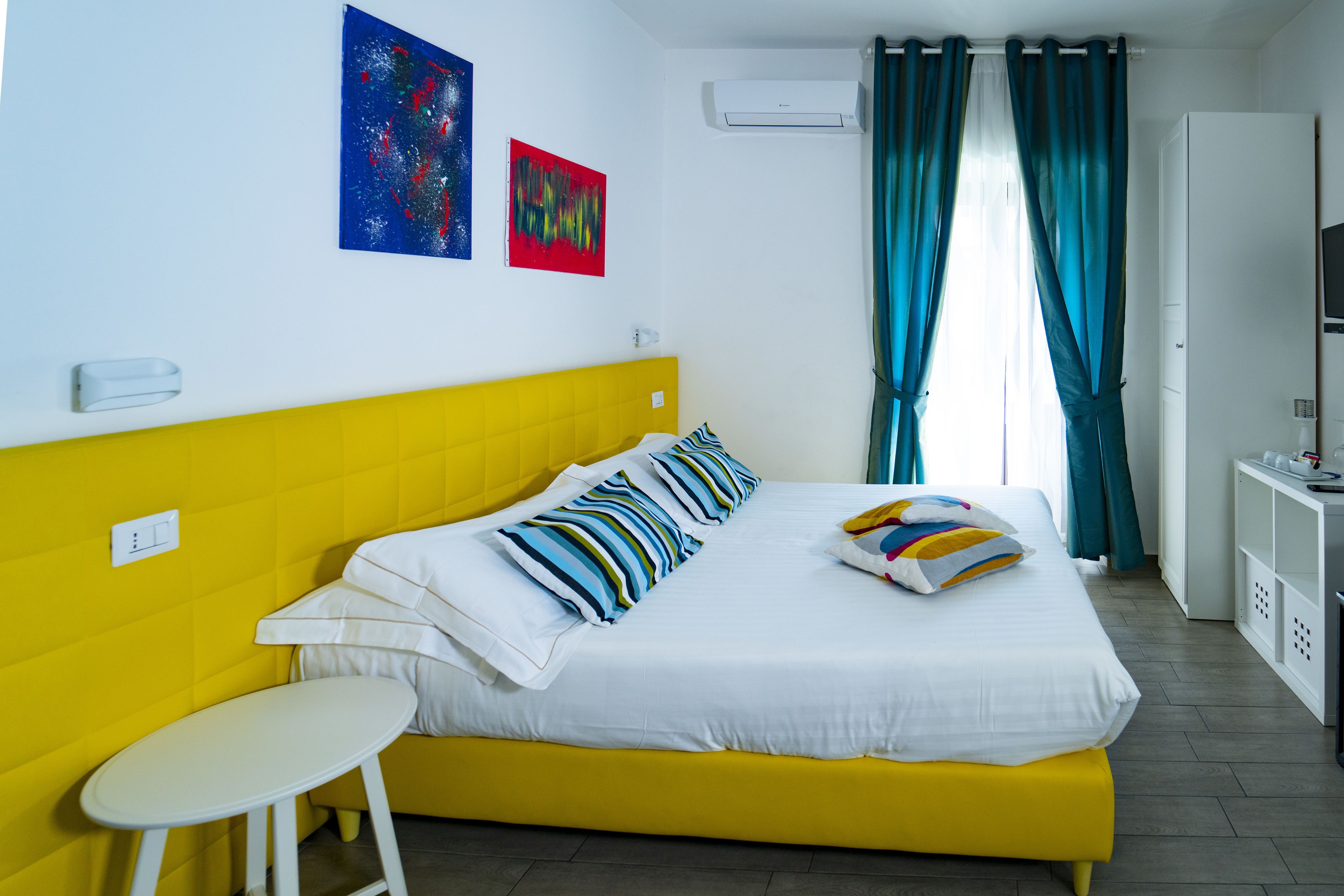 A hotel room with a yellow bed, white pillows, a white bedside table, and a TV on a stand.
