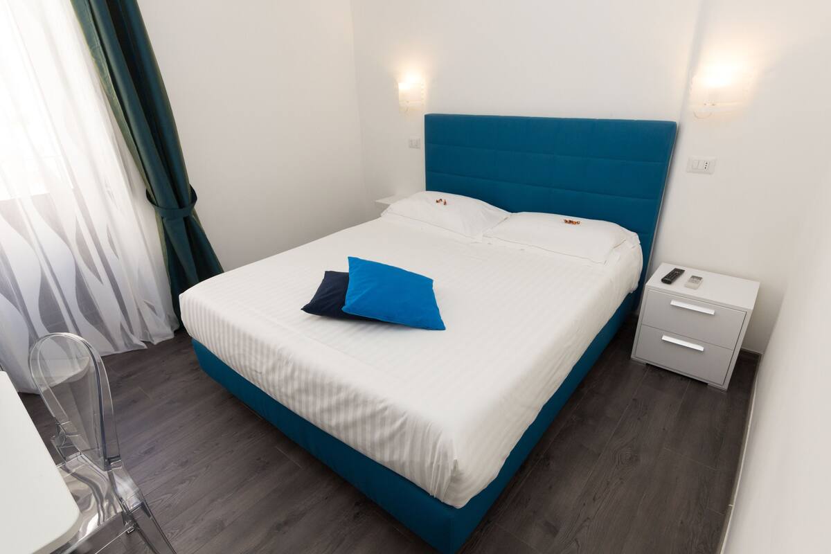 Un letto rifatto con una testiera blu, un comodino con un telecomando e una sedia trasparente.
