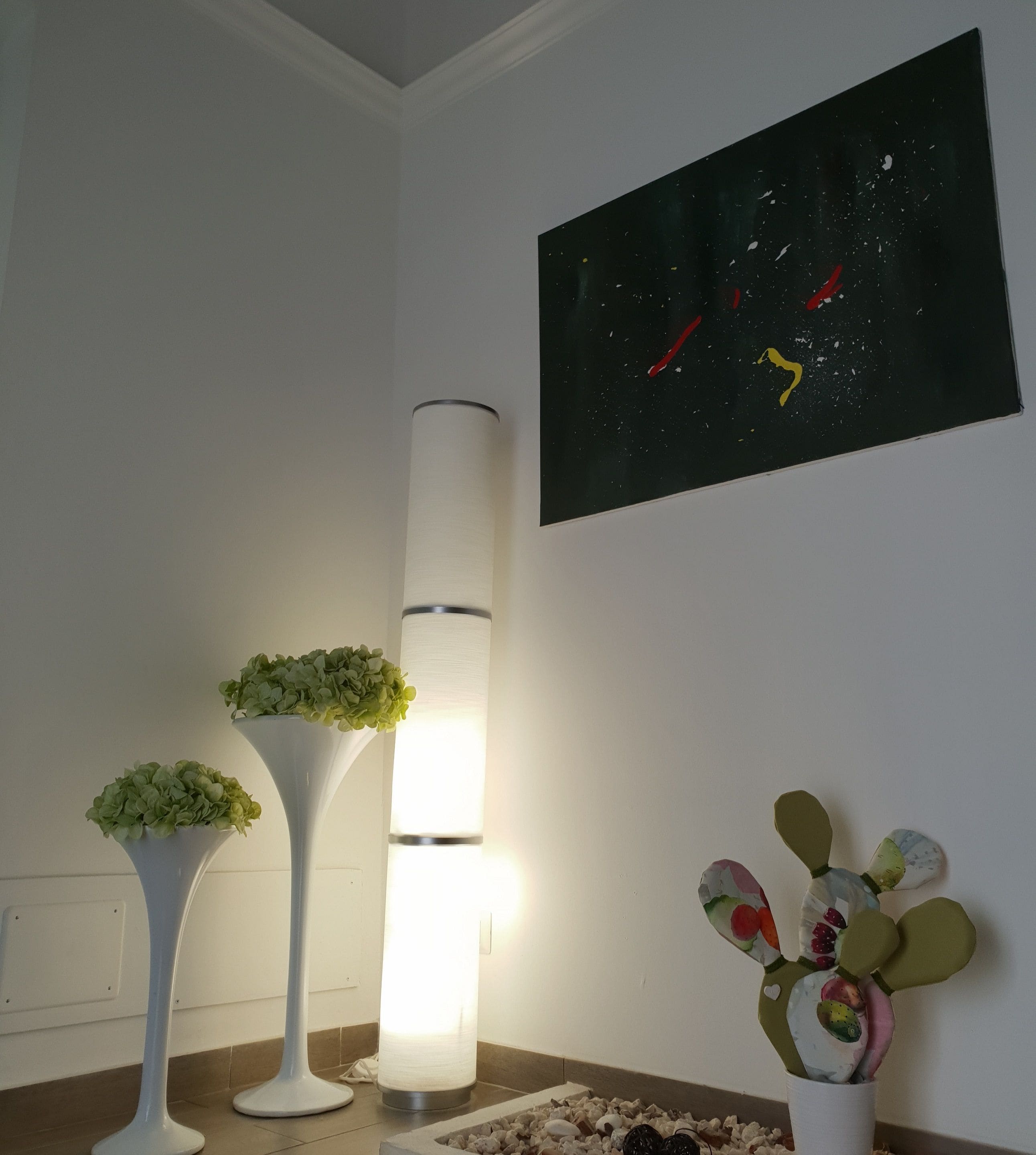 Una habitació amb una làmpada de terra blanca, un vas alt de color blanc amb plantes verdes, i un quadre penjat a la paret.