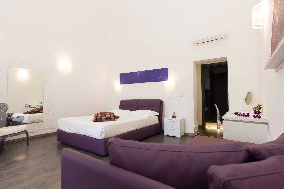 Una camera da letto moderna con un letto viola, pareti bianche e un comodino piccolo bianco.