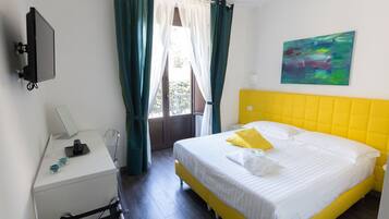 A hotel room with a bed, a desk, a chair, a window with curtains, and a painting on the wall.