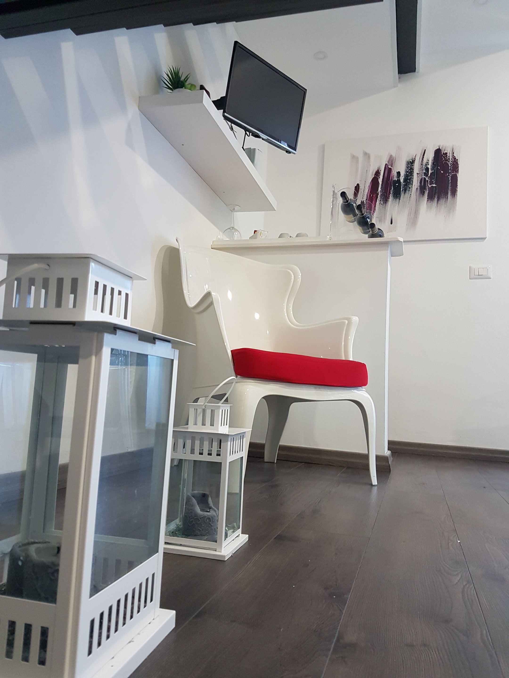 Un interno moderno con una sedia bianca dotata di cuscino rosso, un tavolo bianco e un televisore piatto a parete.
