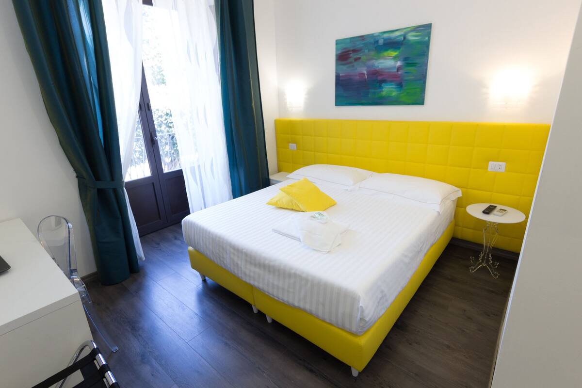 Una camera d'albergo con un letto giallo, tende bianche, un piccolo tavolino rotondo e un quadro appeso al muro.