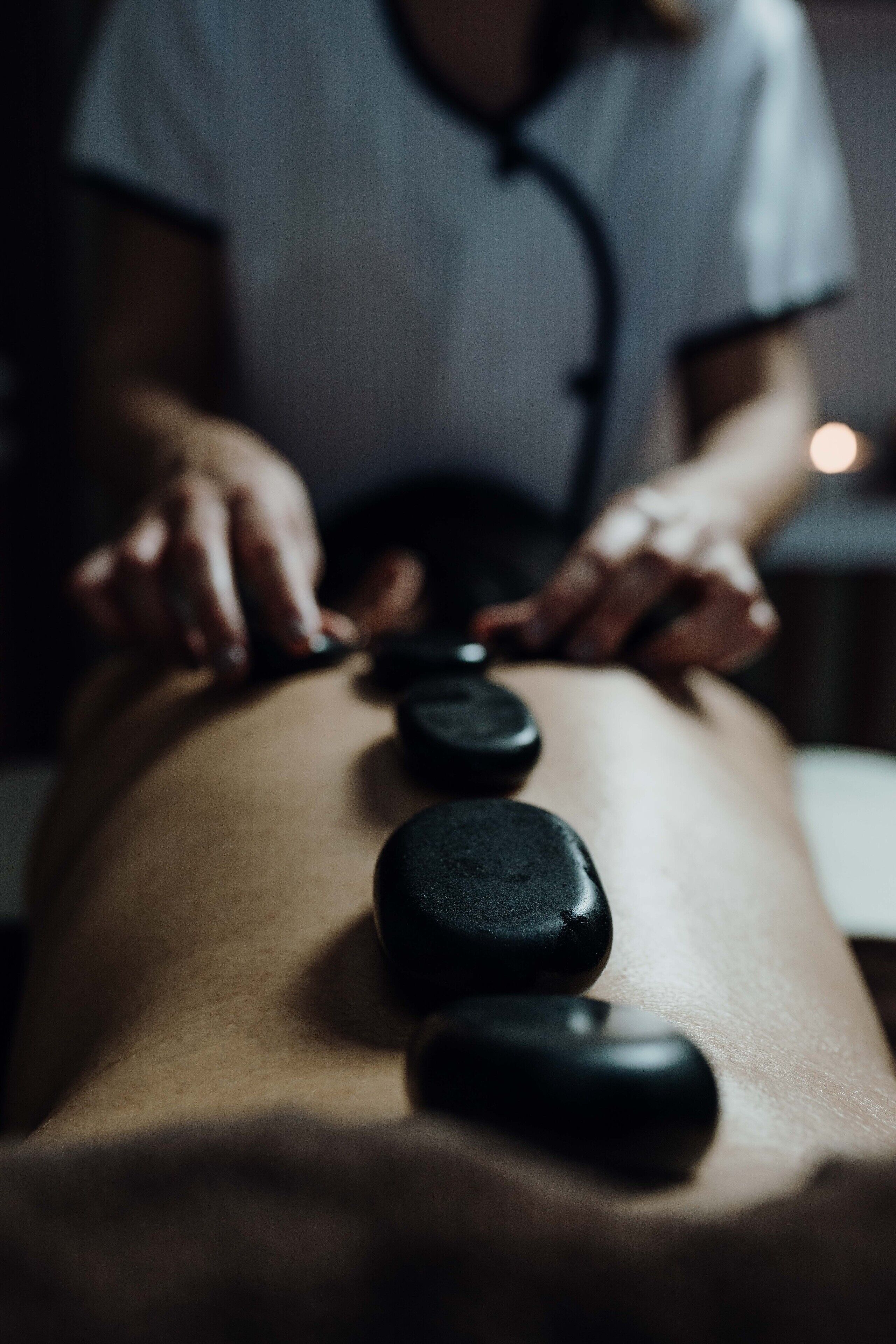 A massage therapy session with a person lying face down and another person applying pressure with stones.