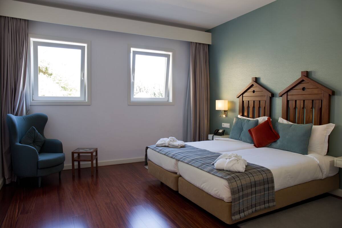 A hotel room with a bed, two windows, a chair, a nightstand, and a lamp.
