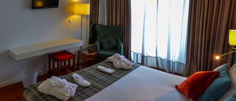 Una habitación de hotel con cama, escritorio, silla, televisor y ventana con cortinas.