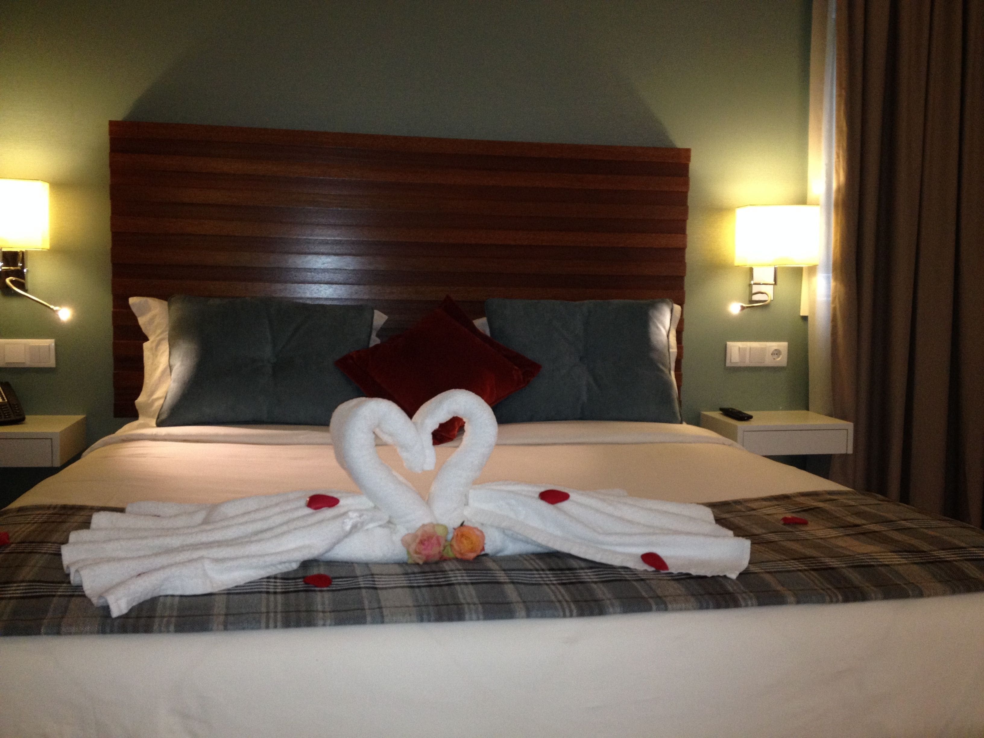 Una cama bien tendida con un arreglo de toallas con forma de corazón, una colcha escocesa y un cabecero de madera.