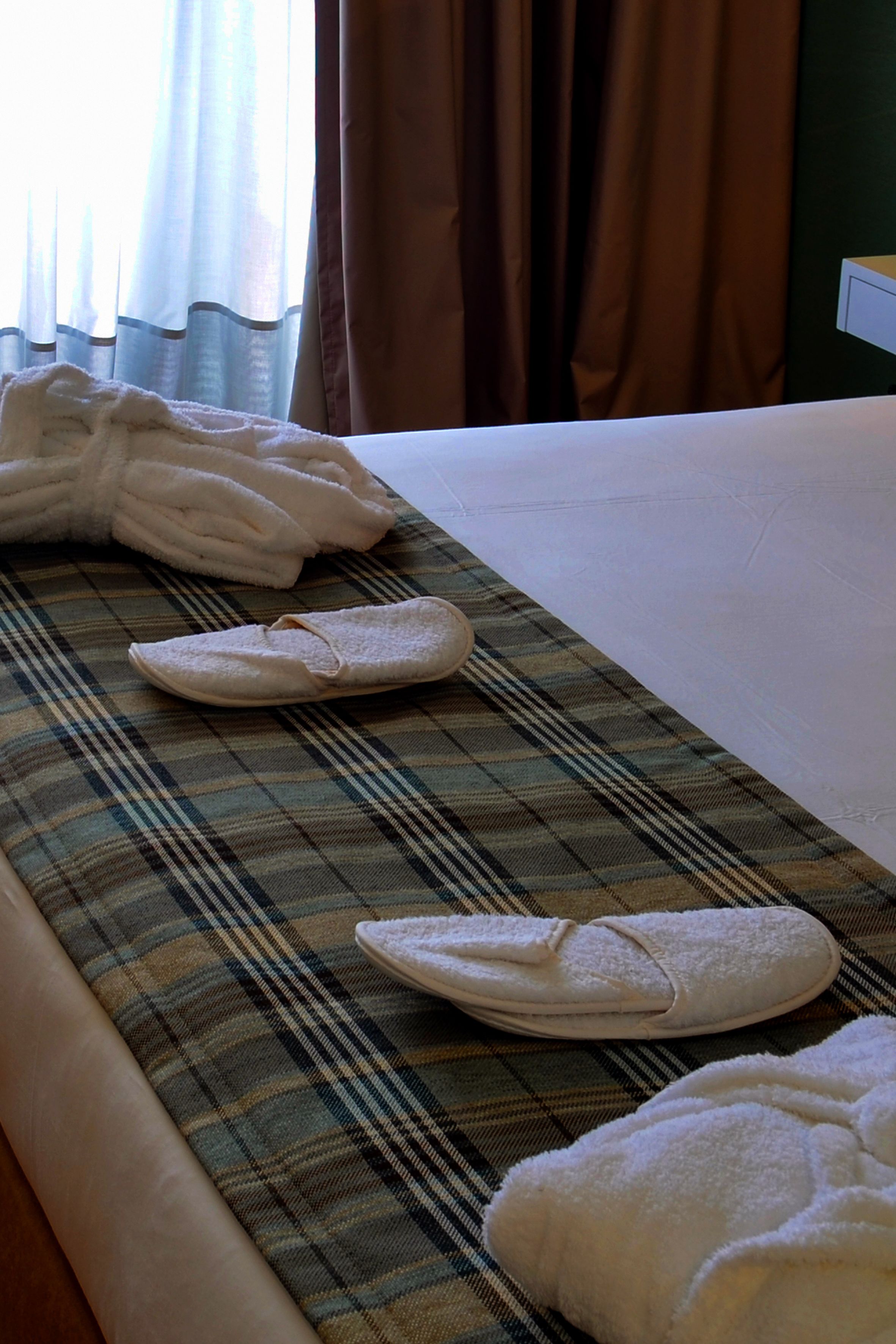 A neatly made bed with a plaid blanket and two pairs of slippers.