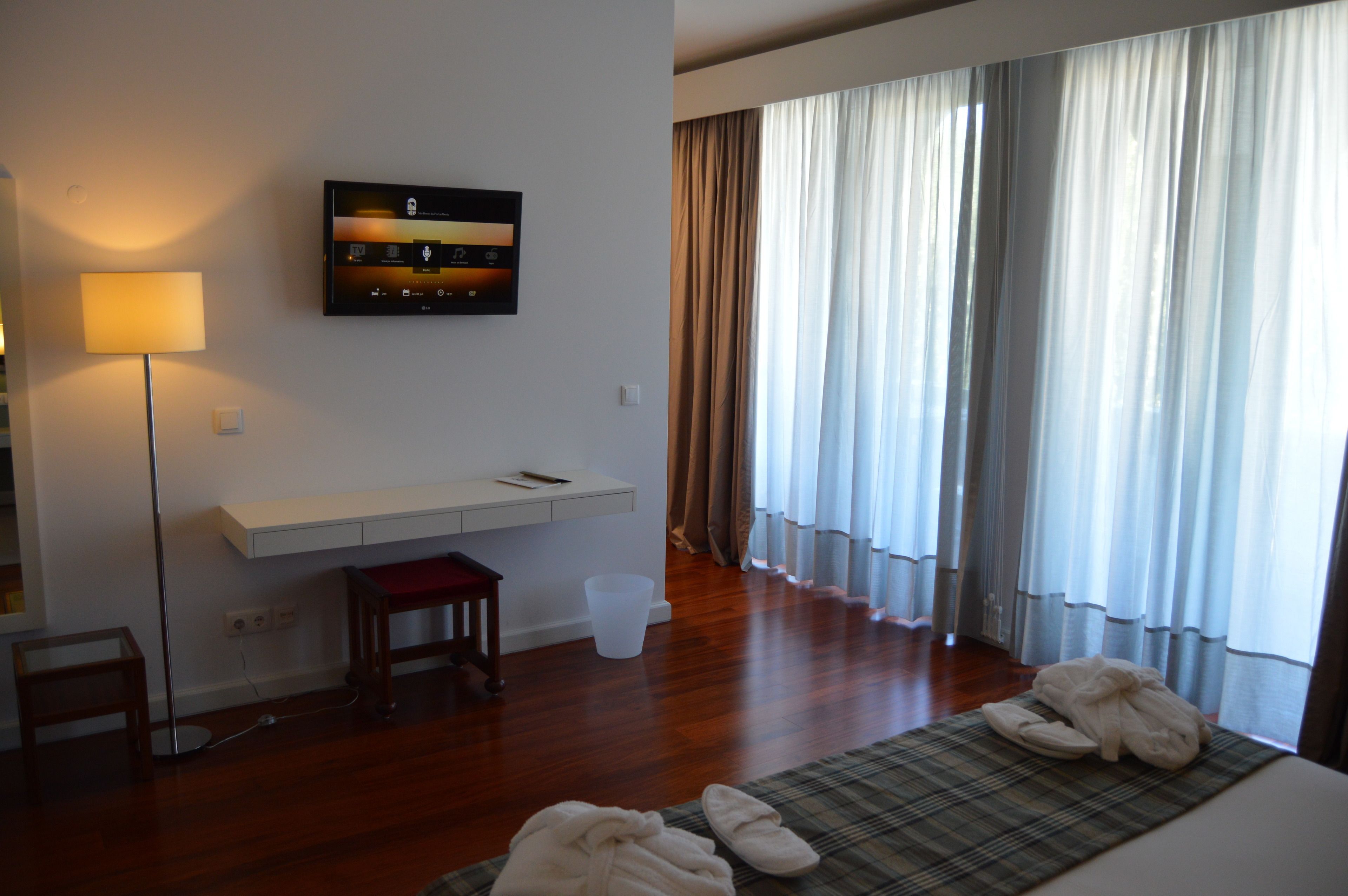 A hotel room with a flat-screen TV mounted on the wall, a desk with a chair, a large window with curtains, and a bed with towels and slippers.