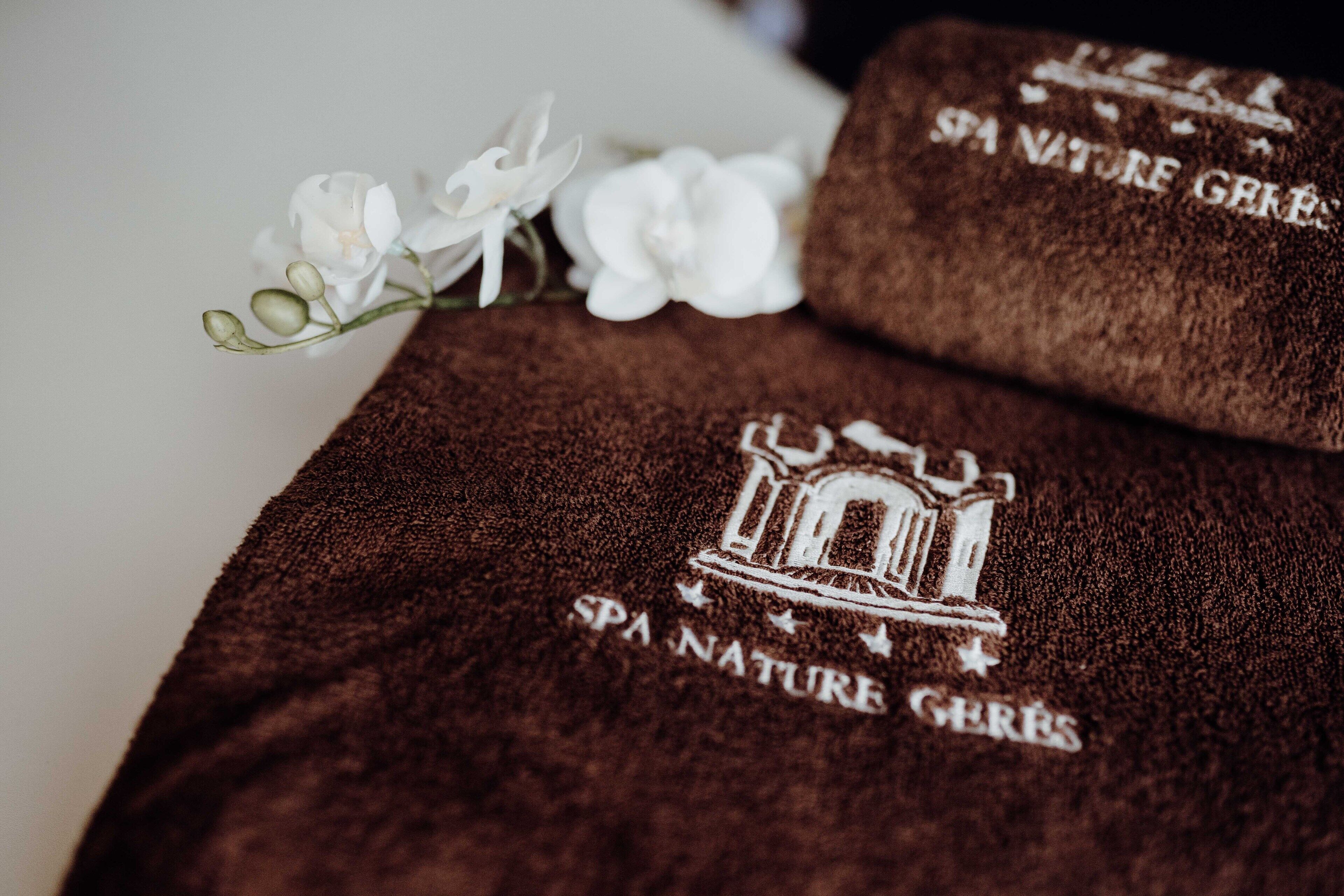 A brown towel with "SPA NATURE GERES" embroidered on it, placed next to a white flower.