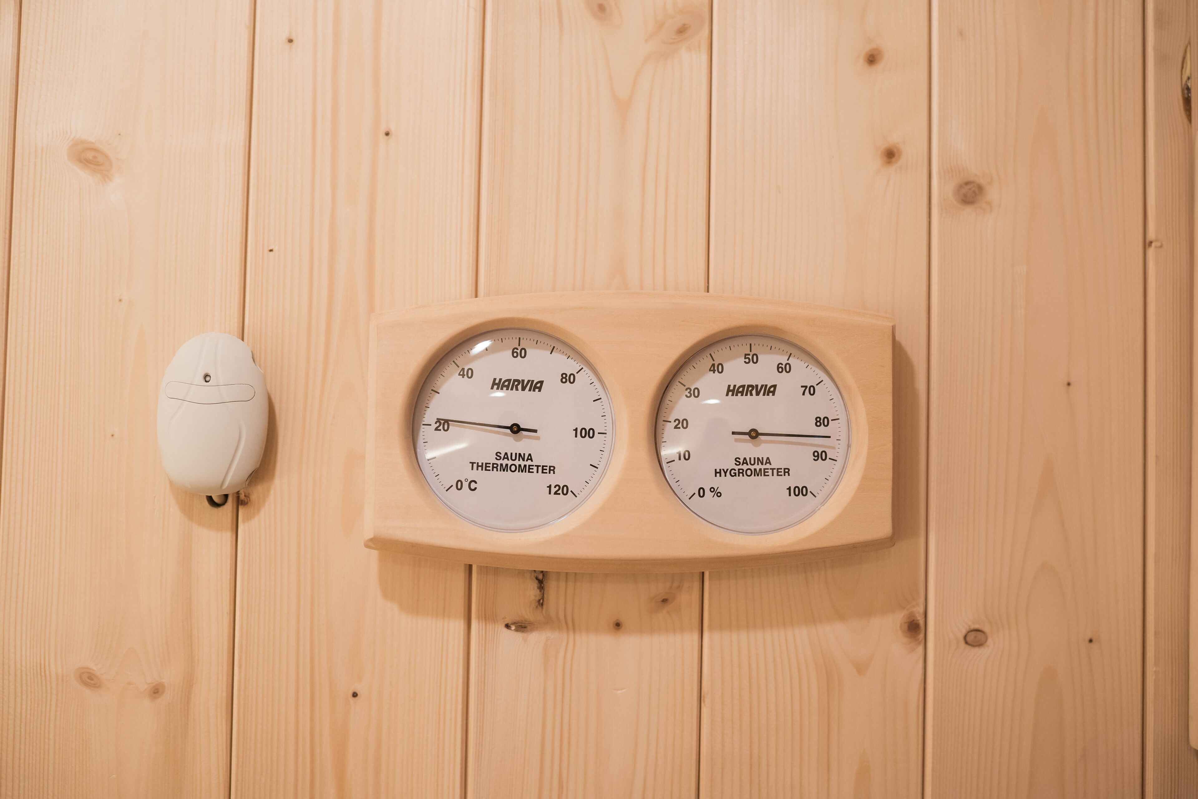 A wooden wall with two circular devices labeled "HARVIA SAUNA THERMOMETER" and "HARVIA SAUNA HYGIOMETER" mounted on it.