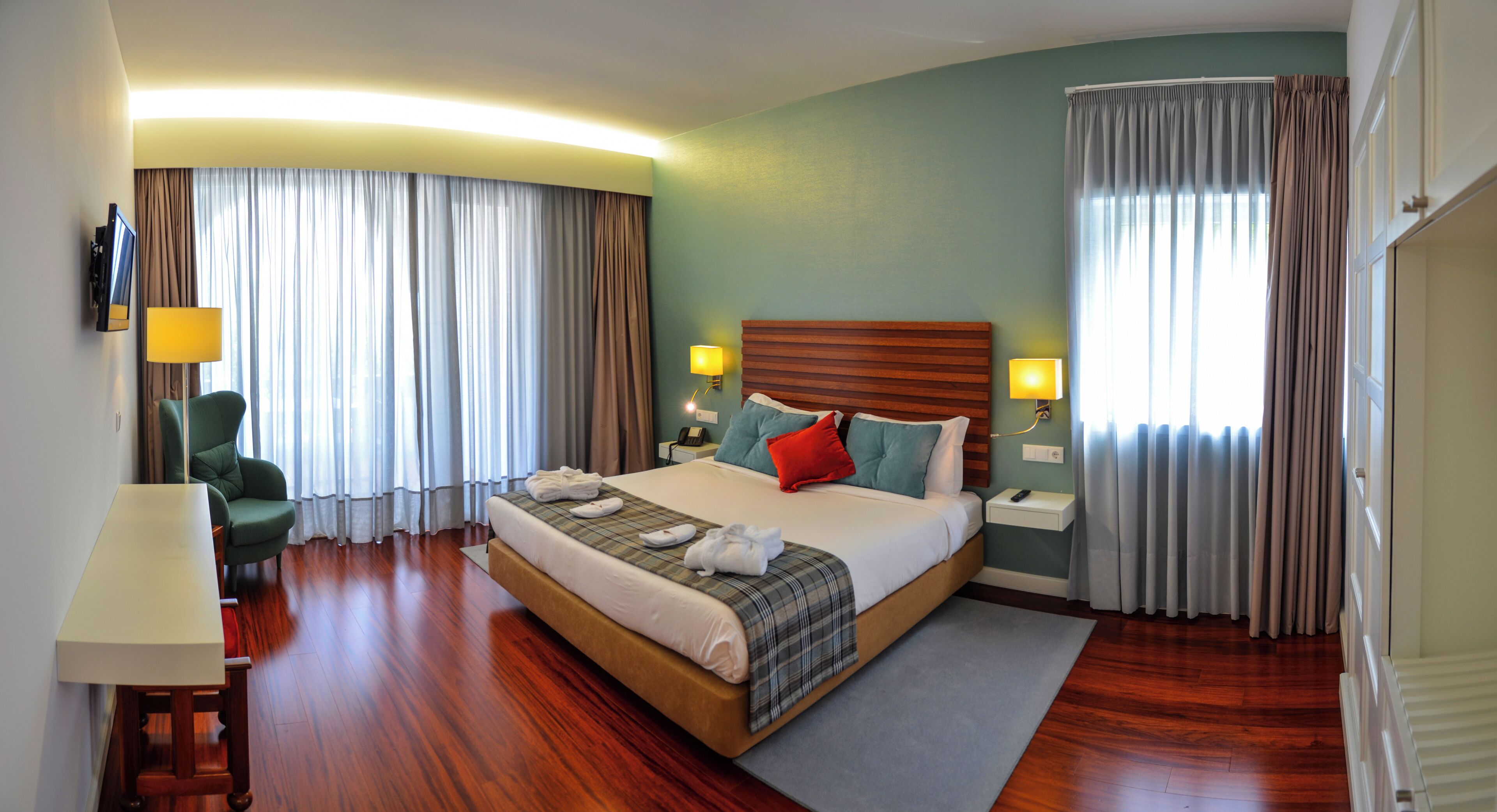 A hotel room with a large bed, a desk, a chair, a TV, and a window with curtains.