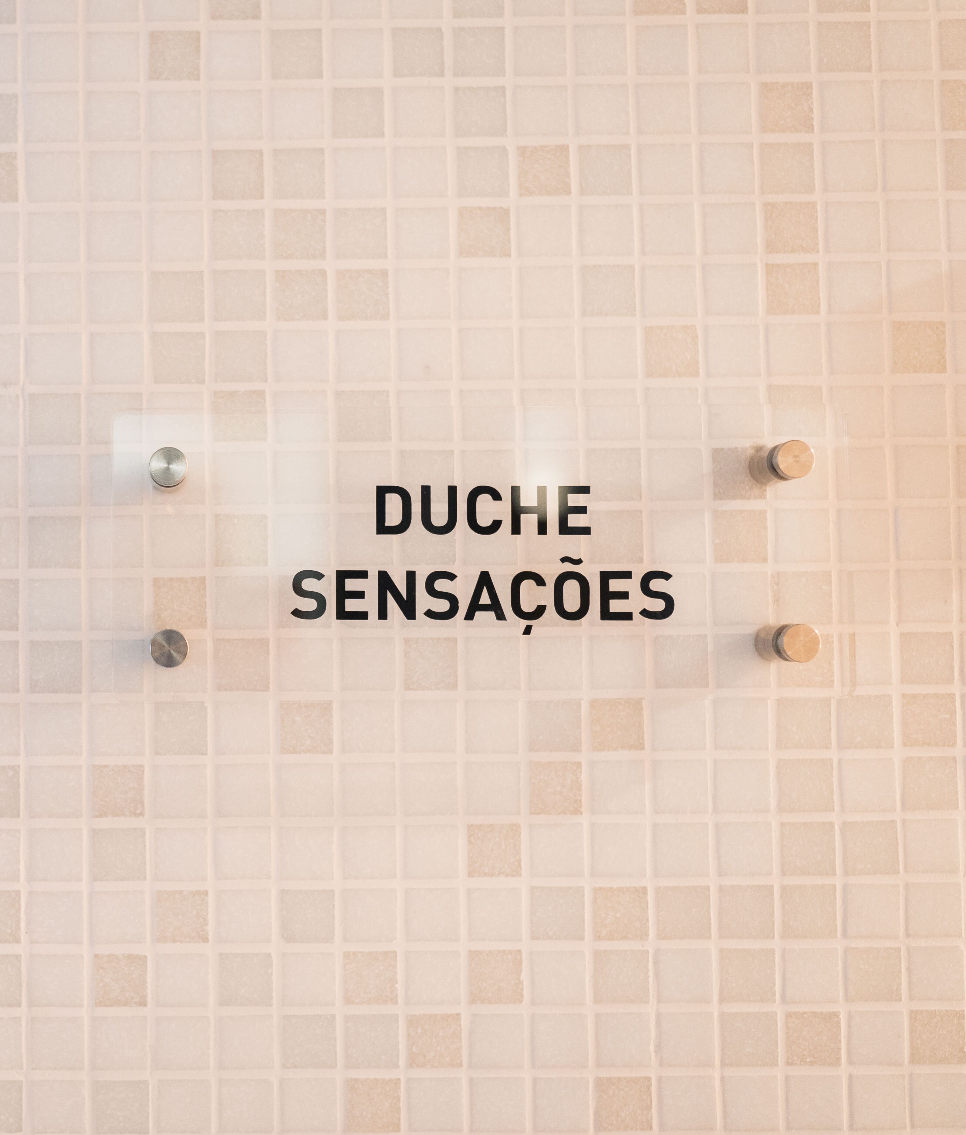 Eine mit Fliesen verkleidete Wand mit dem Text „DUCHE SENSAÇÕES“ und drei sichtbaren Armaturen.