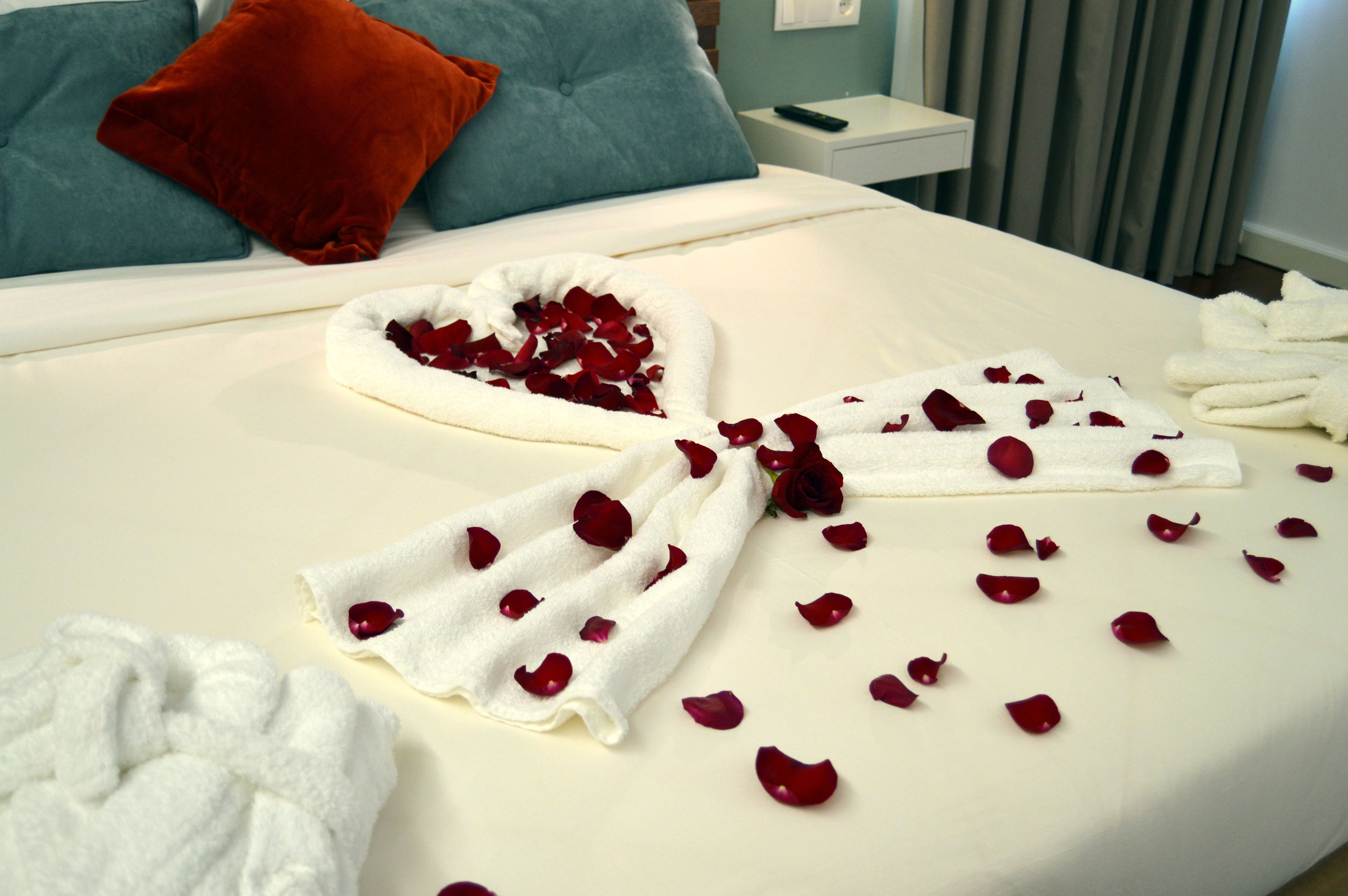 A neatly made bed with rose petals and towels, a red pillow, and a blue pillow on a white headboard.