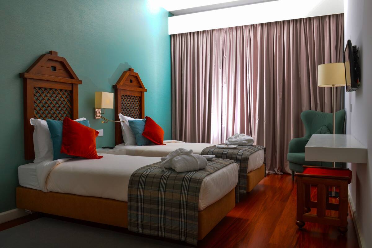 A hotel room with two beds, a green armchair, a small table, and a TV mounted on the wall.