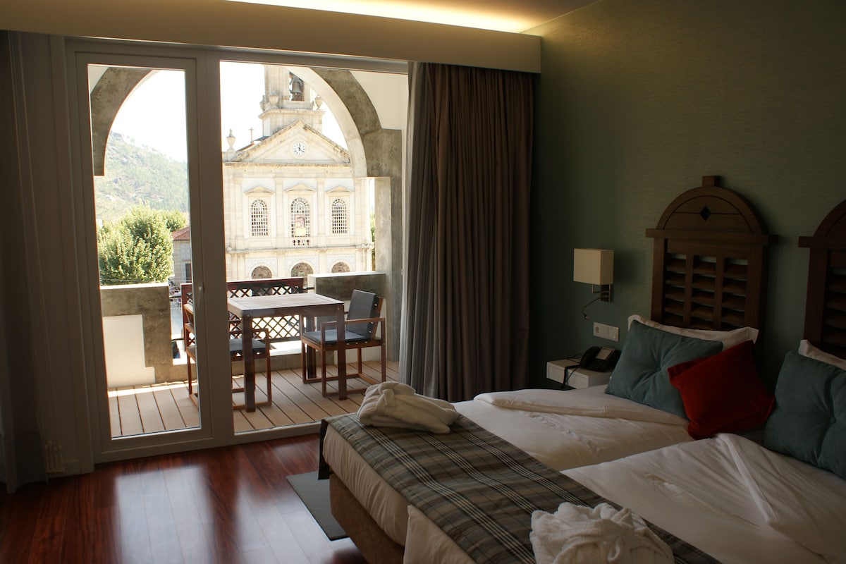 A hotel room with a bed, a desk with a chair, a view of a building through a glass door, and a wooden ceiling.