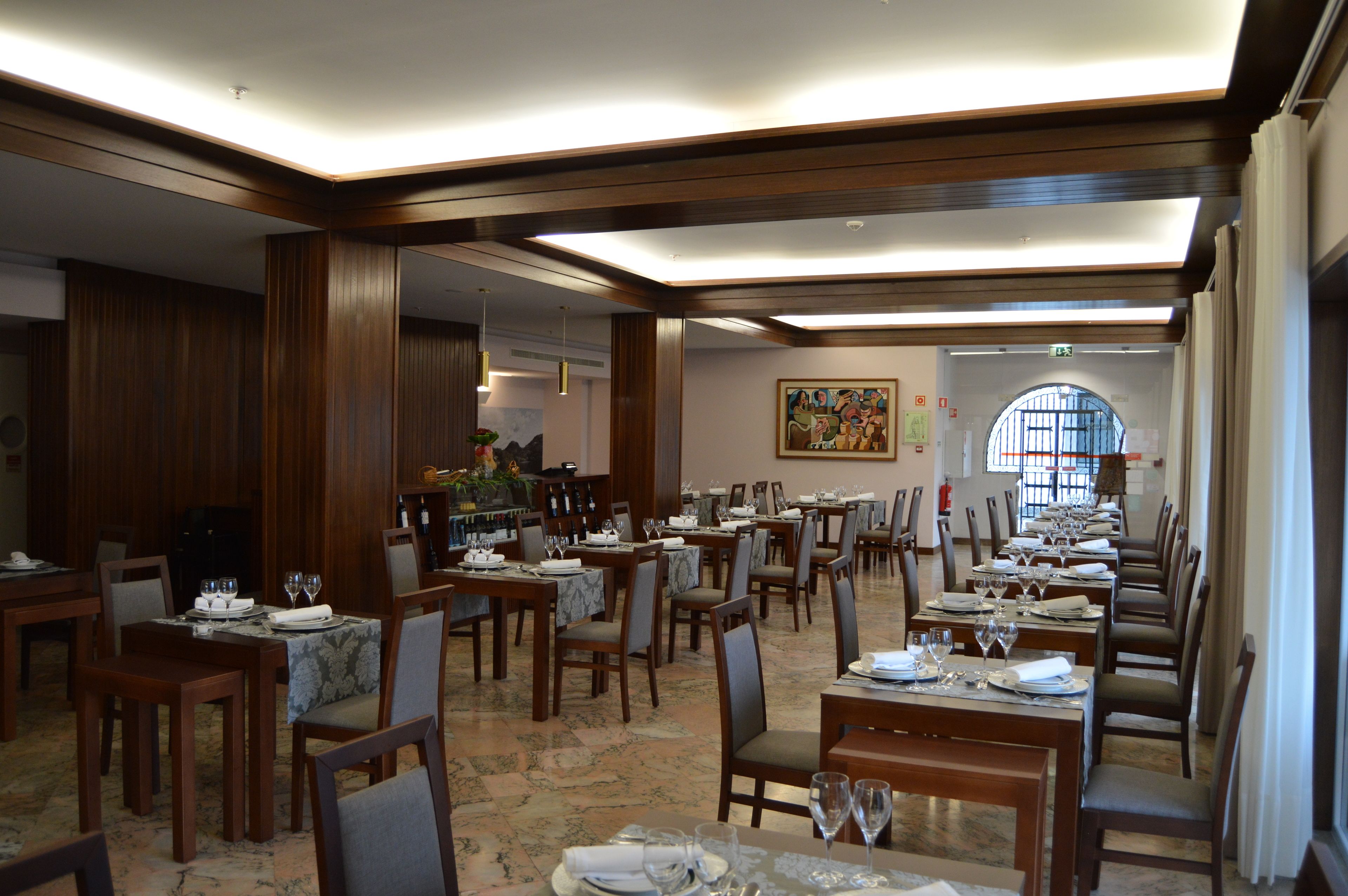 Un vaste espace dédié à la salle à manger, avec plusieurs tables et chaises, un grand tableau accroché au mur et une vue dégagée sur l’extérieur grâce à une fenêtre en arc de cercle.