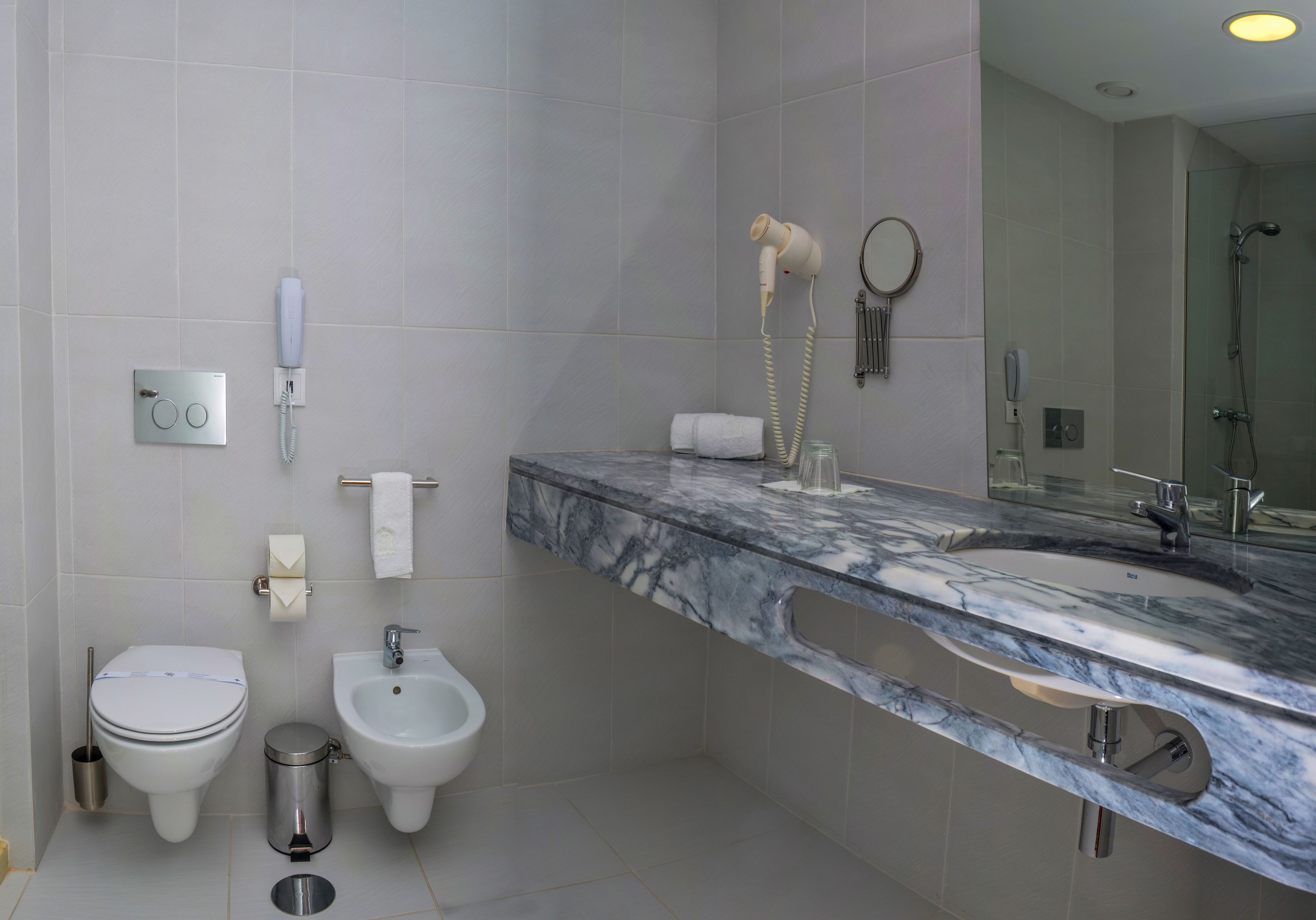 Un baño moderno con lavamanos de mármol, bidé, inodoro y un secador de pelo fijado a la pared.
