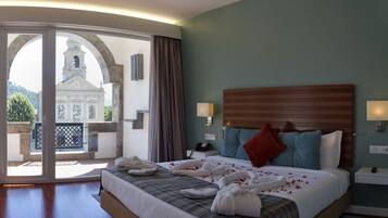 Une chambre d’hôtel avec un grand lit, deux tables de chevet, une tête de lit en bois et une vue sur un immeuble à travers une porte vitrée.