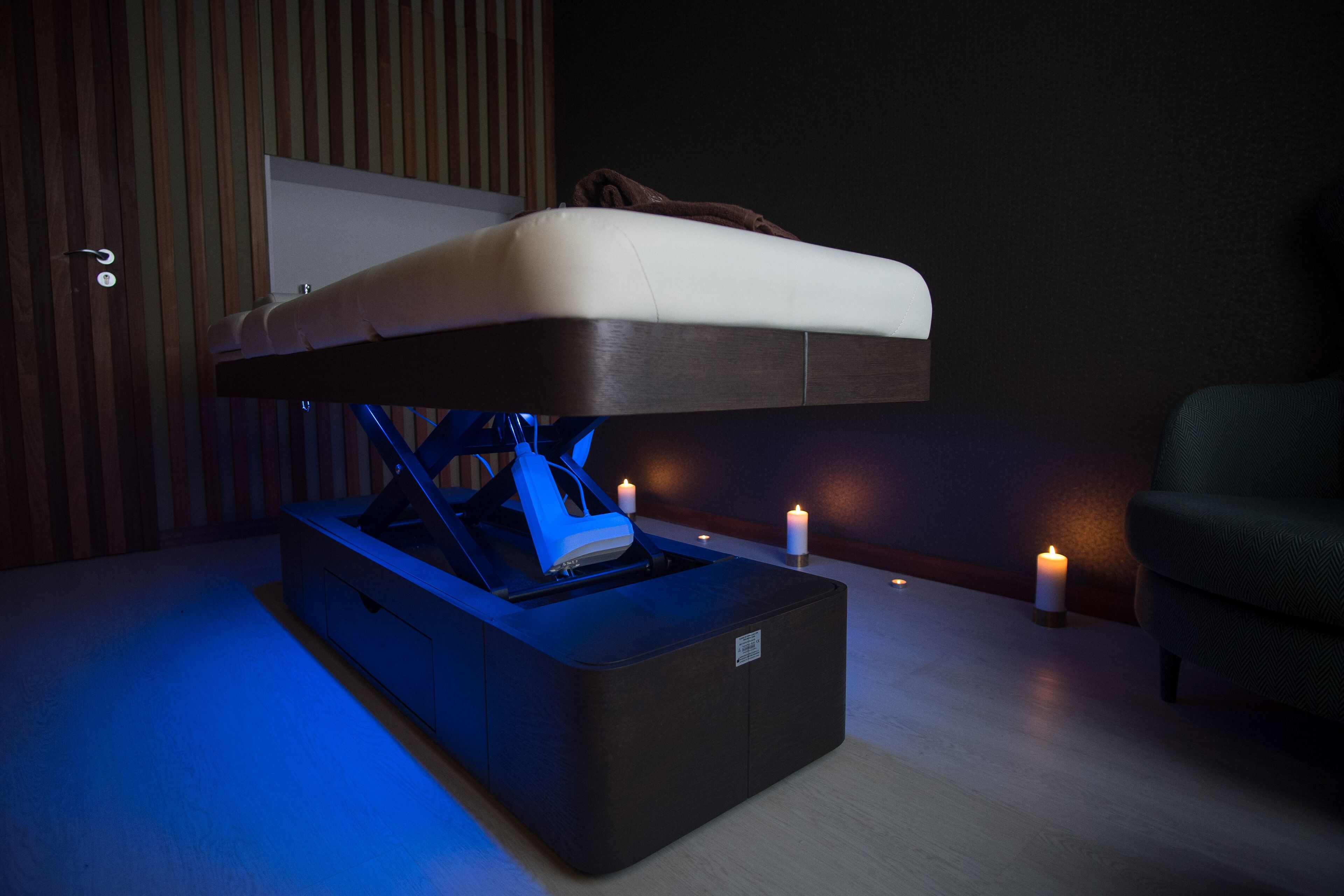 A modern room with a bed elevated on a platform, a massage chair, and candles on the floor.