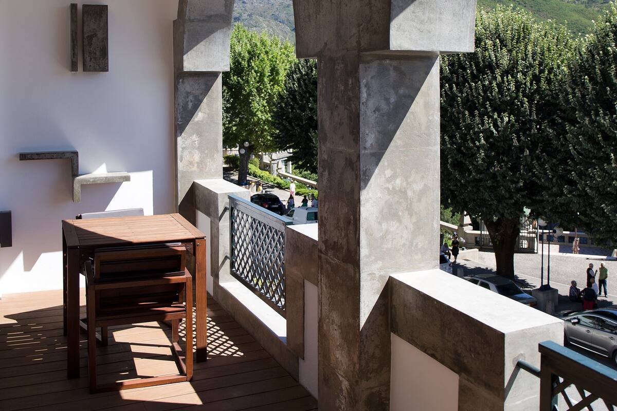 A balcony with a wooden table and chairs, a white wall with a painting, and a view of a tree and hills in the background.