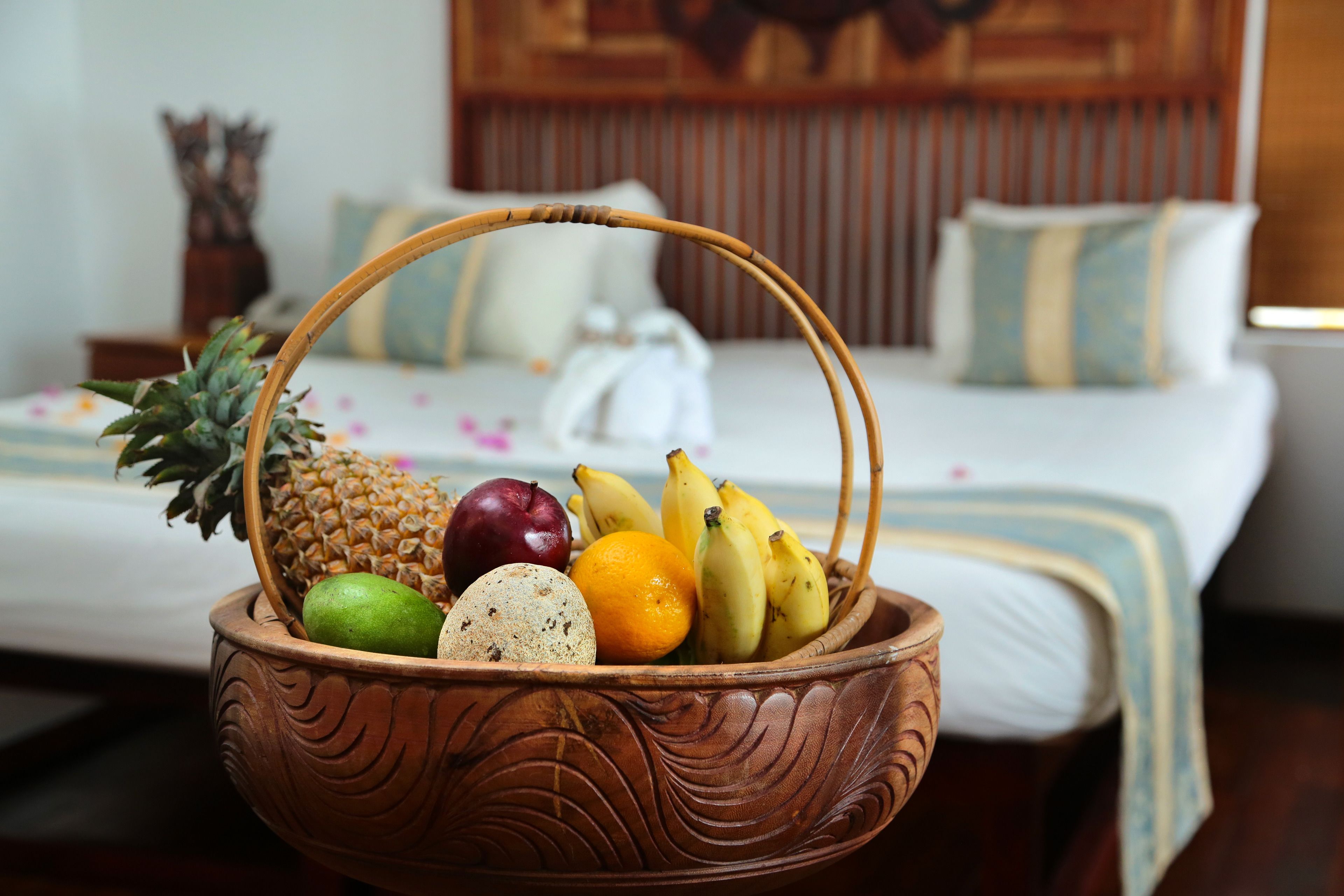 A wicker fruit basket with pineapple, apple, banana, and lime on a bed with white bedding and wooden headboard.
