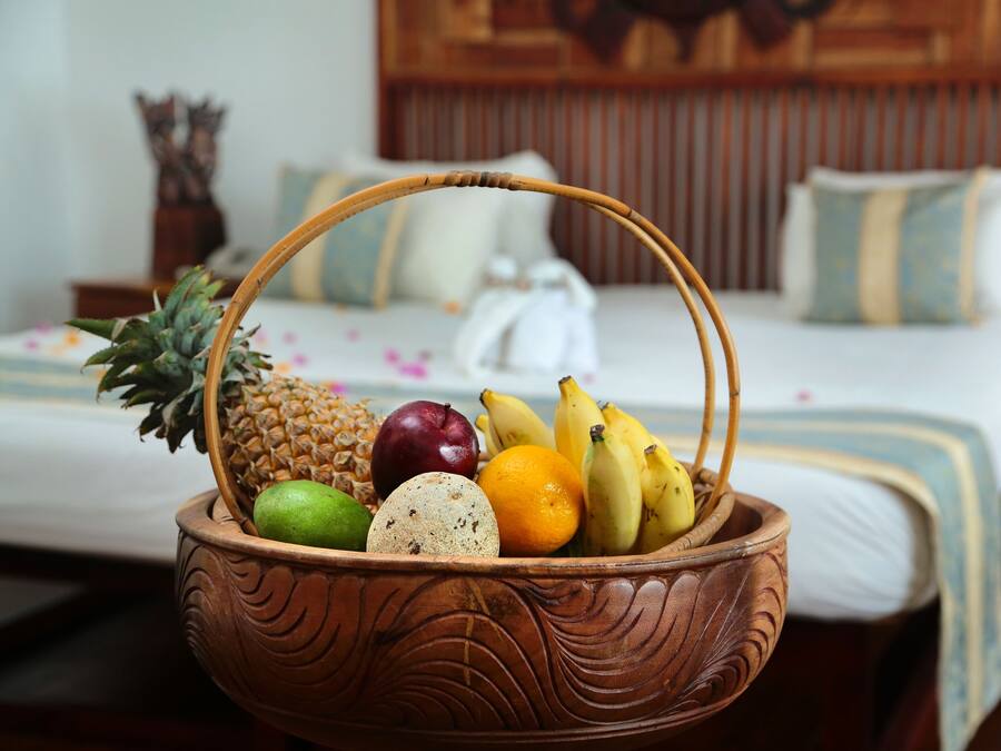A wicker fruit basket with pineapple, apple, banana, and lime on a bed with white bedding and wooden headboard.
