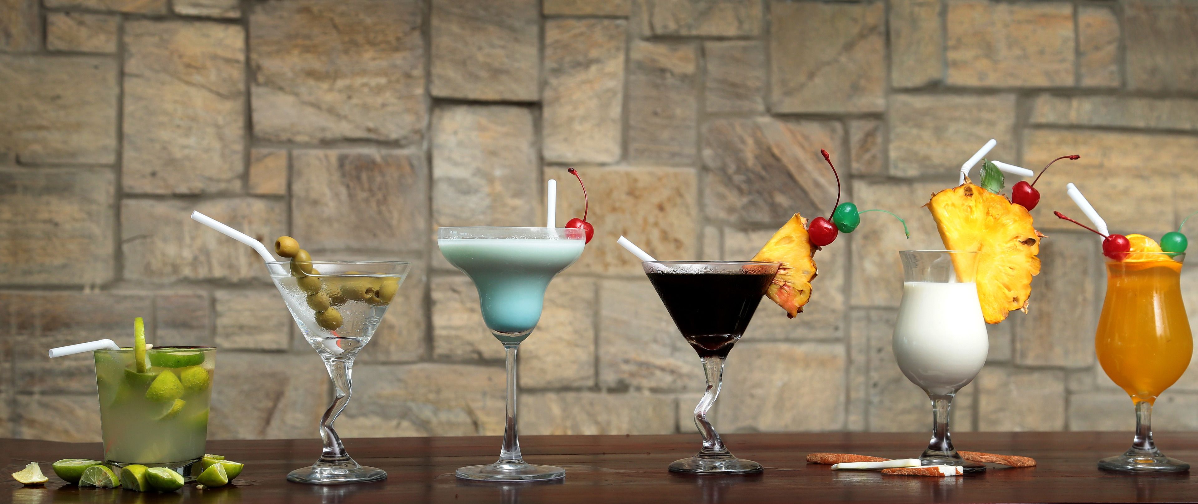 Five cocktails with various garnishes on a bar counter against a stone wall.