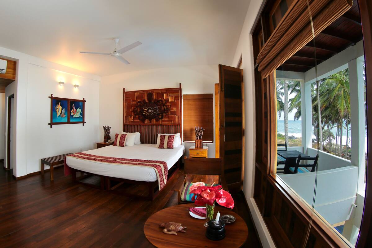 A spacious bedroom with a large bed, wooden flooring, a ceiling fan, and a view of the ocean through a window.