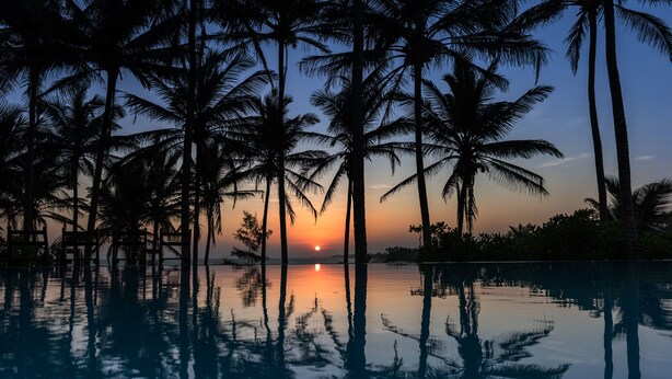 A sunset over a calm pool with palm trees reflected in the water.